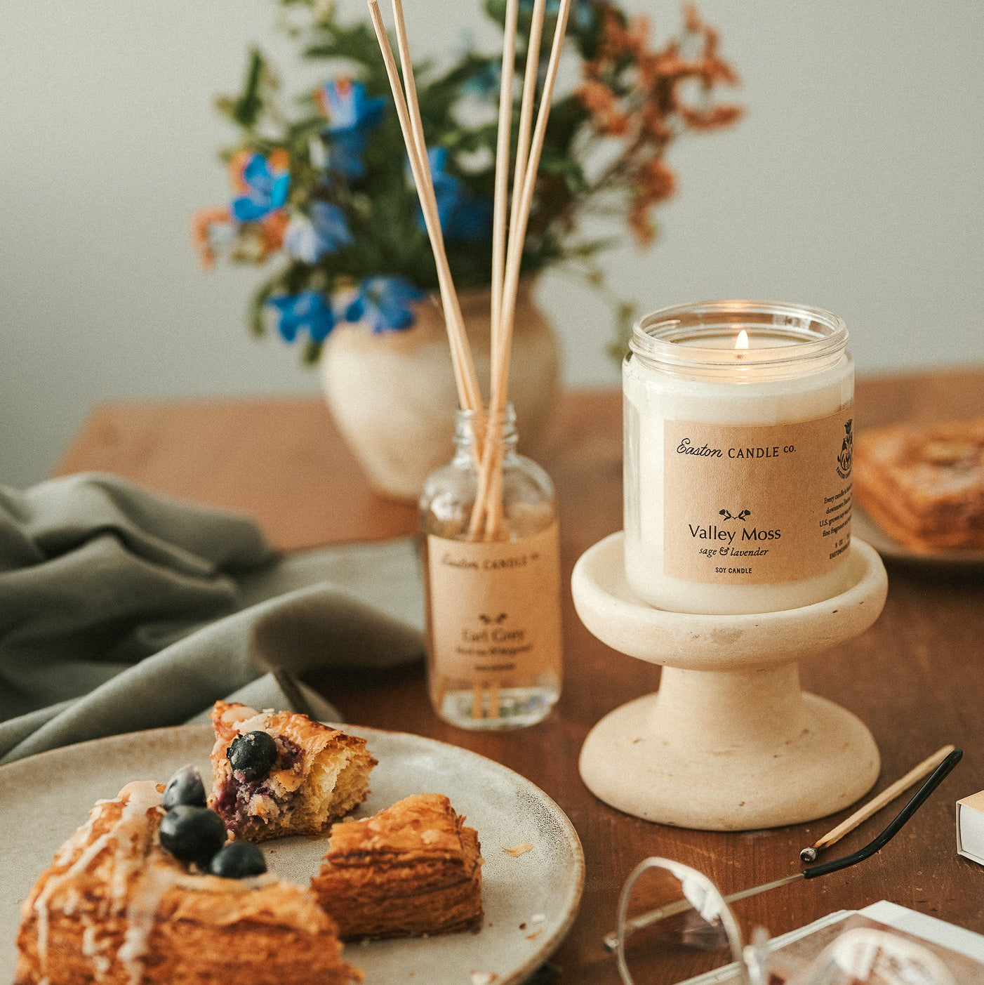Candle on a wooden table with pastries, a diffuser, and flowers in the background